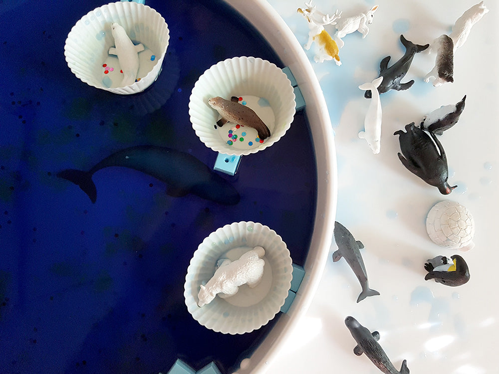 zabawy sensoryczne z galaretką|Miska z niebieską wodą i kawałkami lodu przygotowana do zabawy.|Dziecko wyławiające sitkiem kawałki lodu z niebieskiej wody.|Dziecko wyławiające sitkiem klocki z niebieskiej wody.|Dziecko bawiące się kolorowymi zabawkami ...