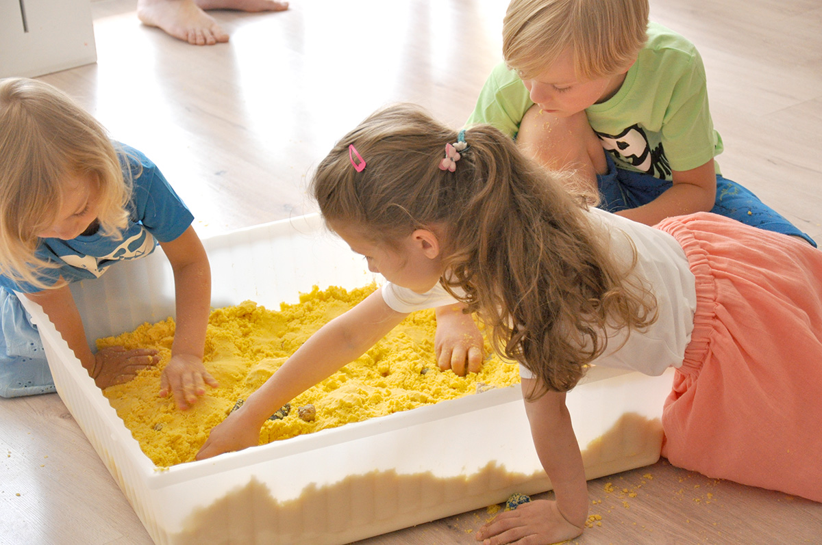 Dzieci zagrzebują zabawki w żółtym piasku kinetycznym.