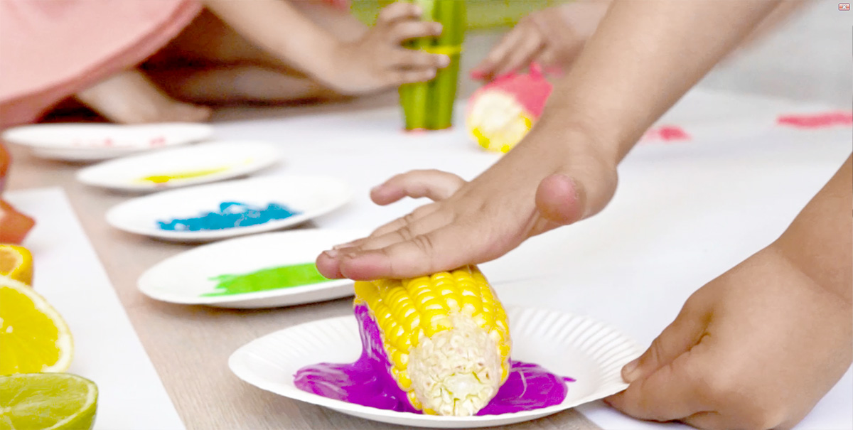 Dziecko maczające kolbę kukurydzy w fioletowej farbie.