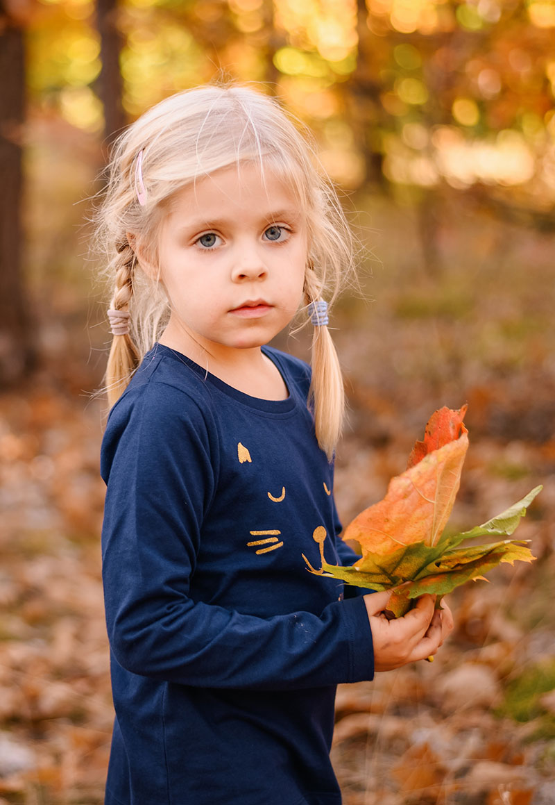 Jesienne zabawy dla dzieci - Mała dziewczynka w granatowej bluzce trzymająca kolorowe liście w ręku.