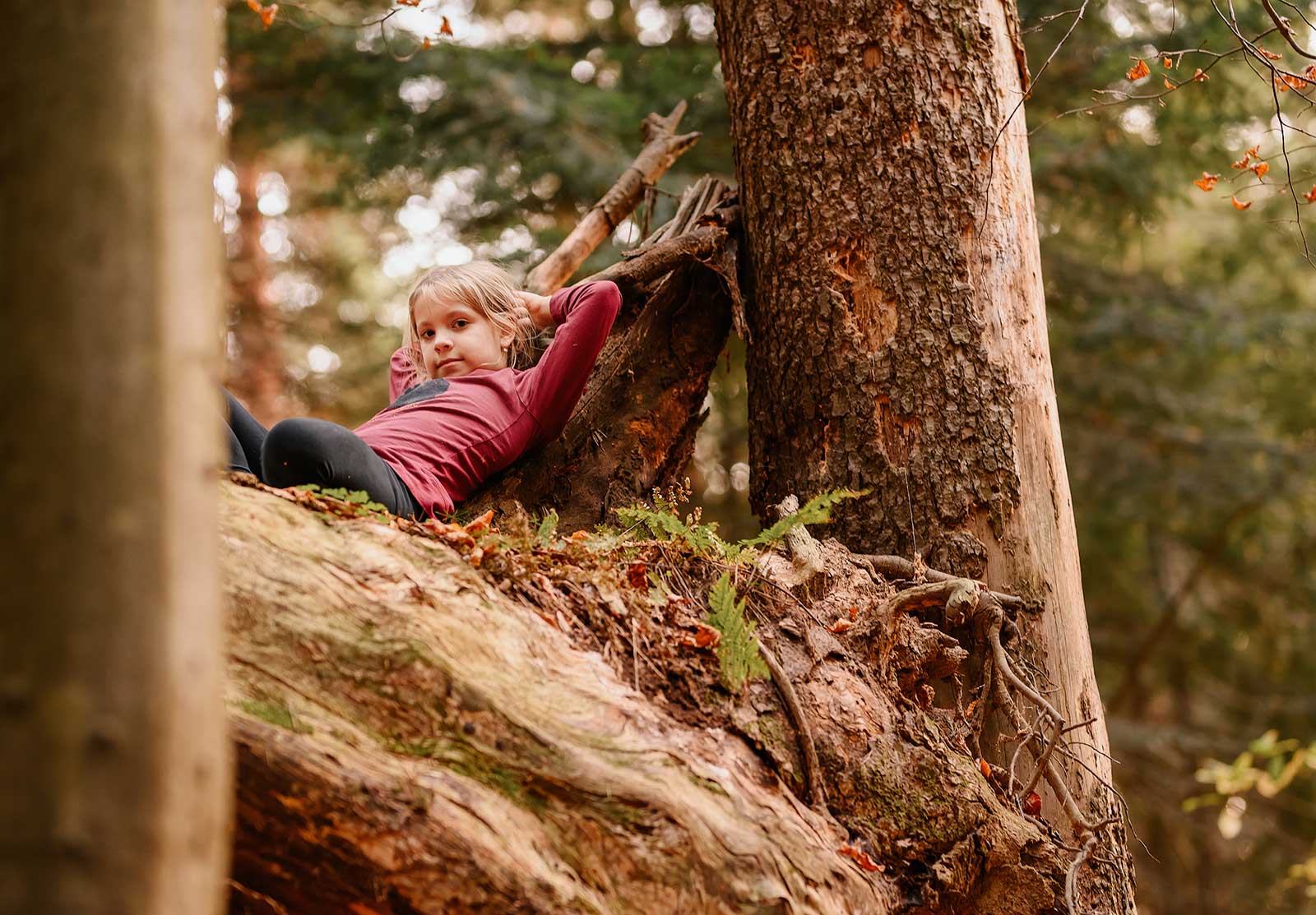 Dziewczynka w bordowej koszulce leży na pniu powalonego drzewa.